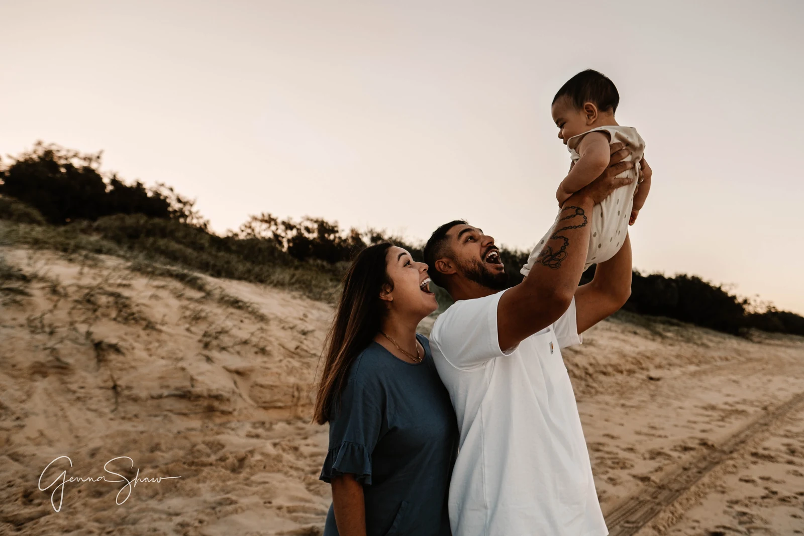 Parents lifting their baby together on the sand dunes at dusk