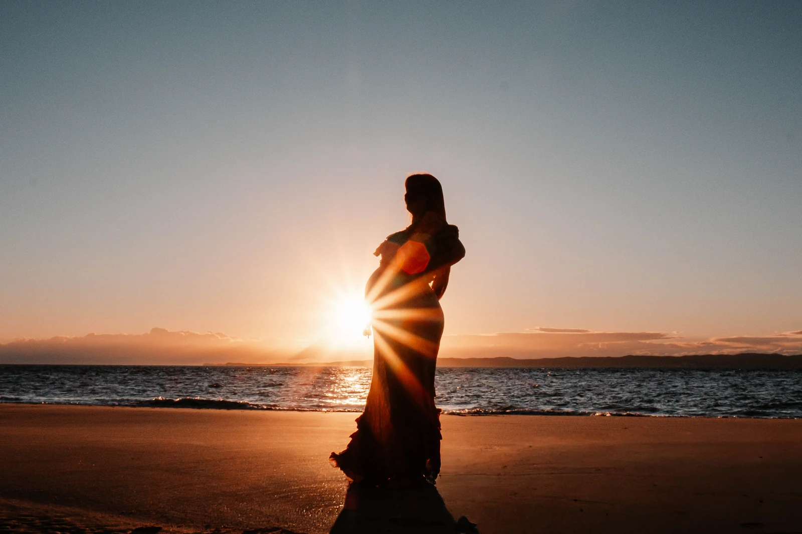Tyler silhouetted on the beach with a sunburst behind her bump